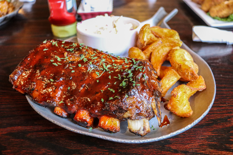 A plate of food on a table, featuring a rack of ribs with a side of potatoes and a small white bowl of sauce or dip.