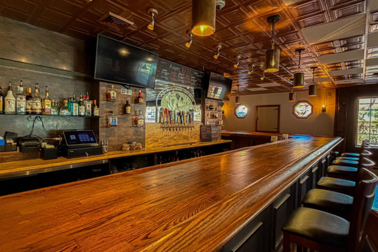A bar with a wooden countertop and chairs, featuring a TV, bottles of liquor, and a clock on the wall.