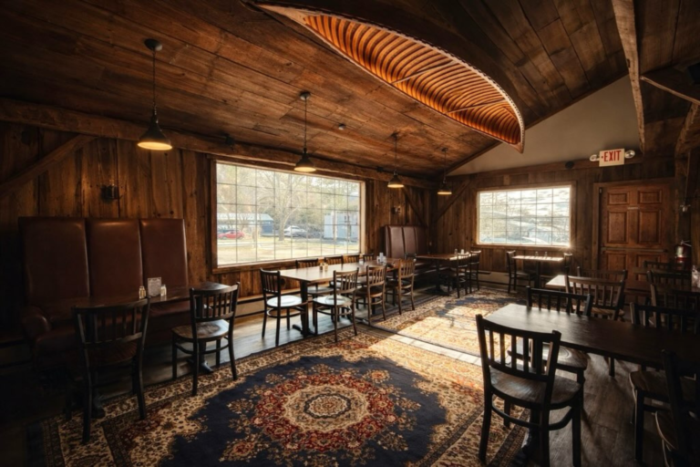 A restaurant dining room with wooden walls and ceiling, featuring a canoe-shaped light fixture, wooden tables and chairs, and a large window.