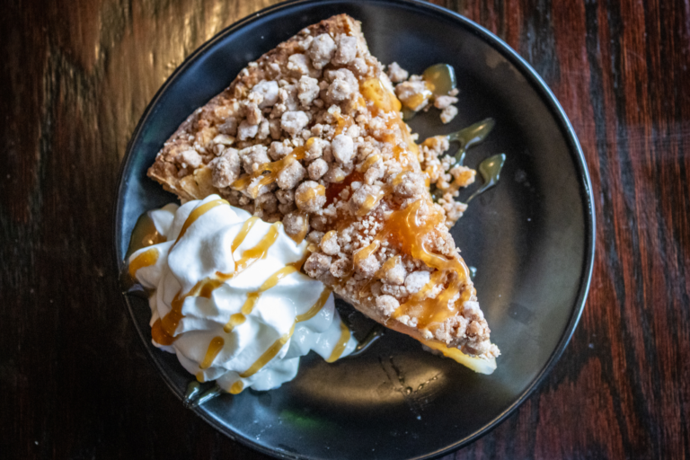 A slice of caramel apple pie with whipped cream on a black plate, set against a dark wood table.