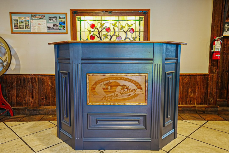 A wooden reception desk with a stained glass window featuring apples above it, situated in a restaurant with a tile floor and wooden paneling on the walls.