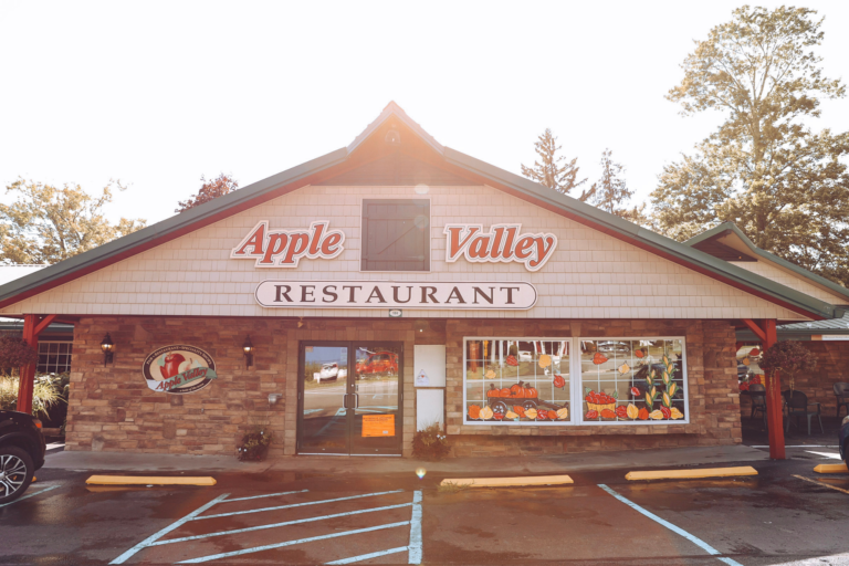 The image depicts a restaurant building with a parking lot in front, featuring a sign that reads "Apple Valley Restaurant" and a large window displaying a vibrant display of pumpkins and gourds. The building's exterior is constructed from brown brick, complemented by a green roof and a white sign with red lettering. A car is parked on the left side of the building, while a tree is visible in the background.