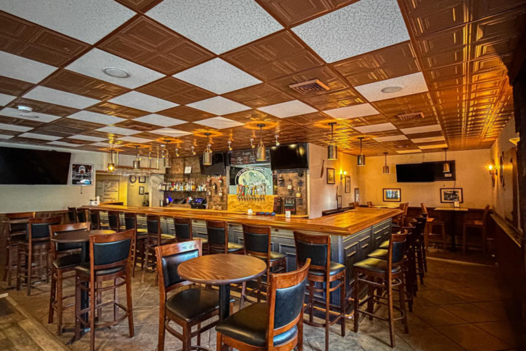 A dimly lit bar with a wooden bar top, featuring a flat-screen TV, a selection of bottles and glasses, and a few chairs and tables scattered around. The ceiling is made up of square tiles in a checkerboard pattern, adding to the cozy ambiance of the space.