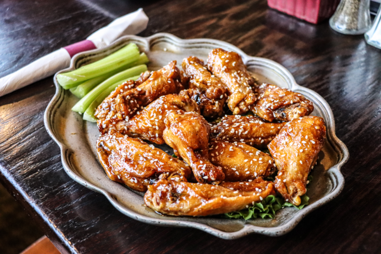 A plate of chicken wings with sesame seeds and a side of celery on a dark wooden table.